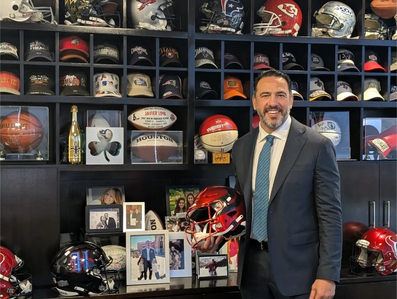 Javier Loya holding a red football helmet in front of shelves of signed helmets, caps and game balls
